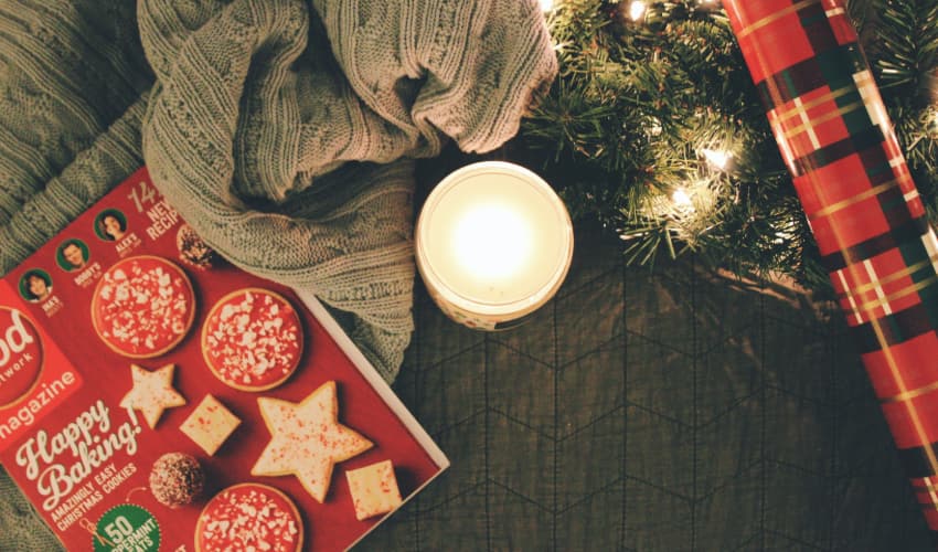 a lit candle among a spread of festive items: a holiday cookie recipe book, a knit blanket, a pine bough strung with lights, and red plaid wrapping paper