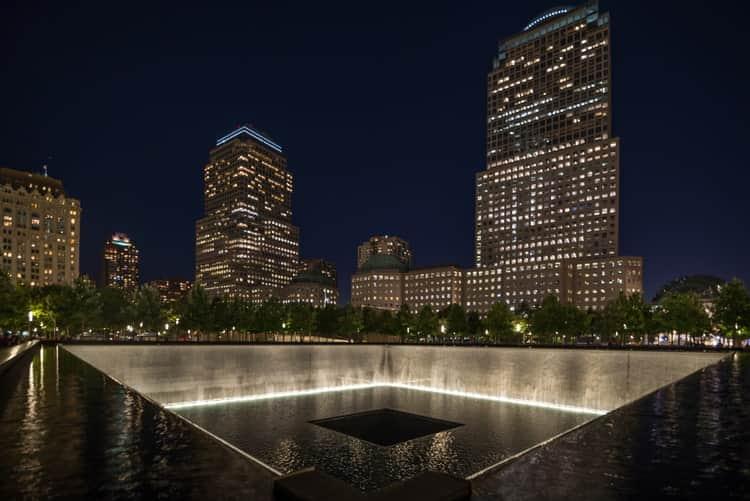 reflecting pool at the 9/11 memorial shines at night with city lights in the background