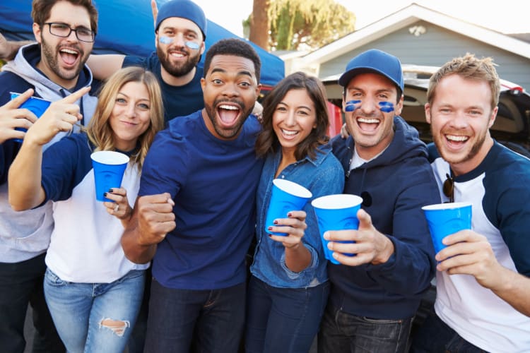A group of fans dressed in blue hold cups and cheer at a tailgate