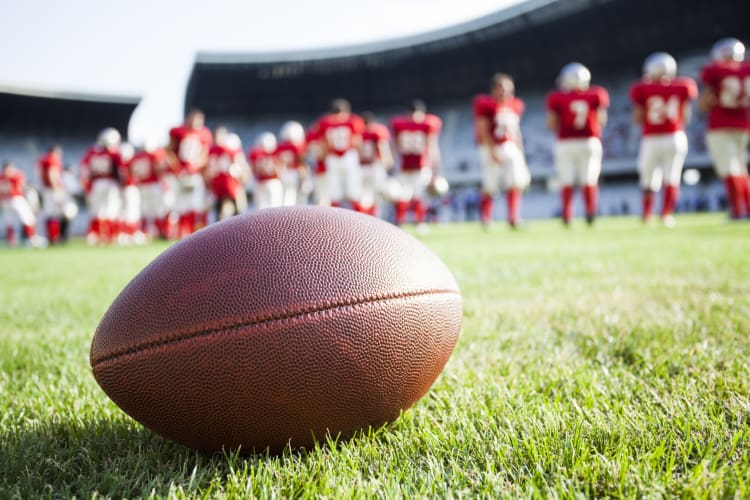 a lone football on a stadium field, with football players and bleachers visible int he background