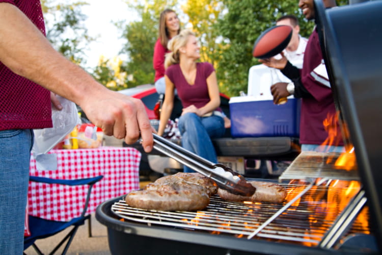 a fan grills meats at a tailgate, other fans visible chatting in the background