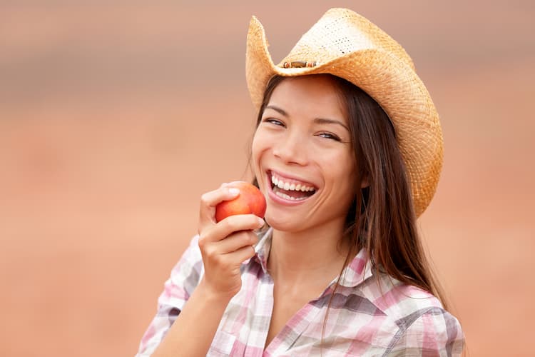 Woman wearing a cowboy hat smiles before biting into a Texas peach.