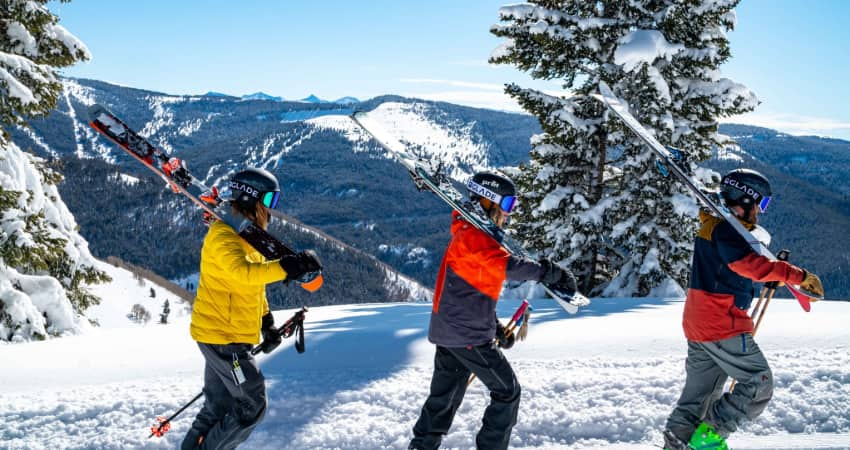 three skiers walk along a mountain ridge with their skis and poles over their shoulders