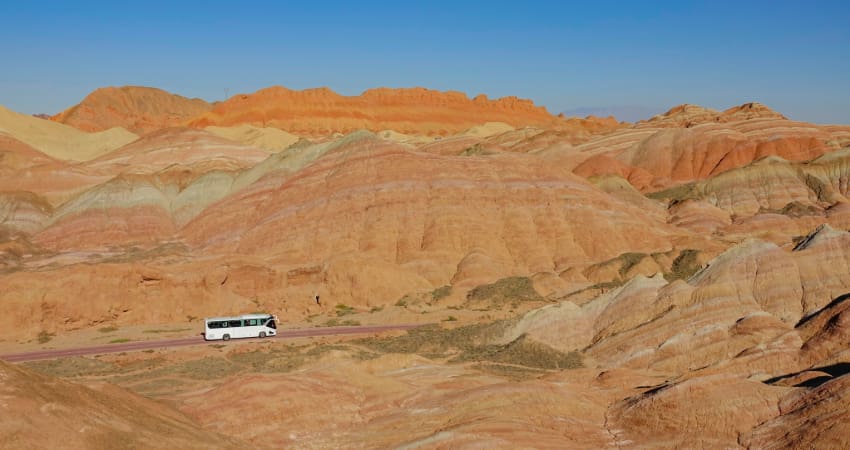 A lone charter bus drives through the desert, rock formations in the distance