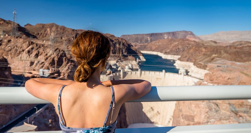 A lone woman stands at an overlook railing, looking down on the Hoover Dam and Lake Mead