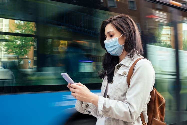 Woman waiting on bus with phone and mask