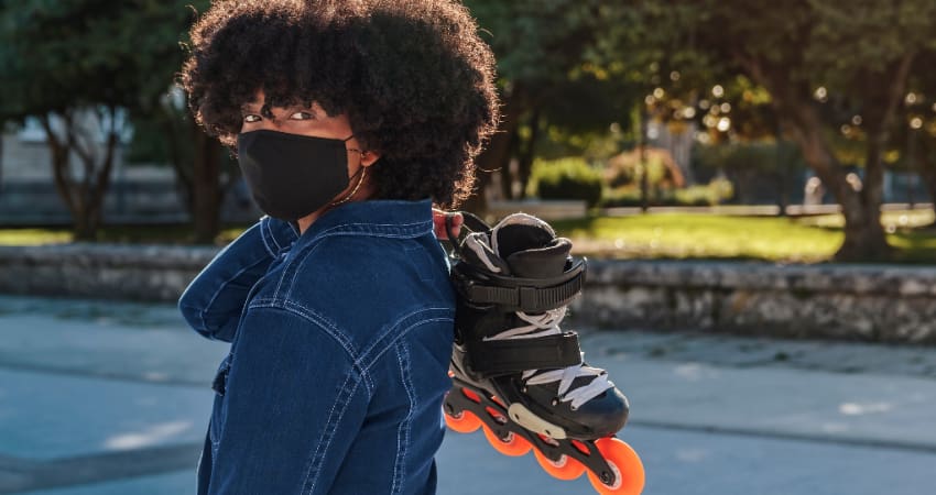 A woman wearing a mask carries a pair of rollerblades in a green public park