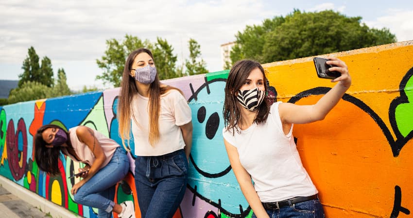 Three friends wearing face coverings pose for a selfie in front of a bright outdoor mural