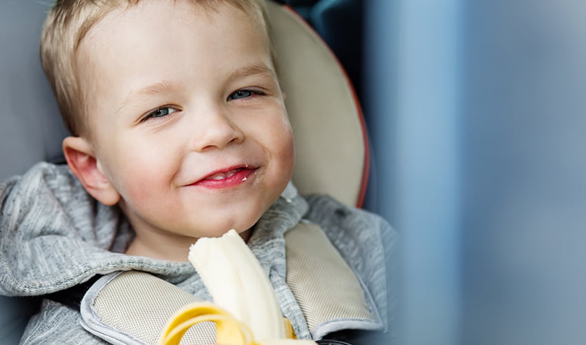 A child eats a banana and smiles from his seat in a vehicle