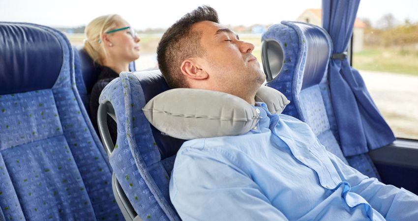 Man sleeping on the bus with a neck pillow while a fellow passenger sleeps behind him.