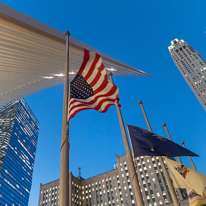 A flagpole rises above the New York City streets, skyscrapers visible behind it