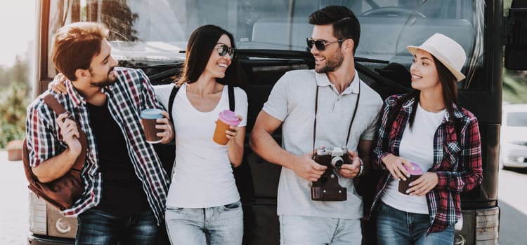 four friends stand in front of a bus while holding drinks and cameras, all smiling at each other