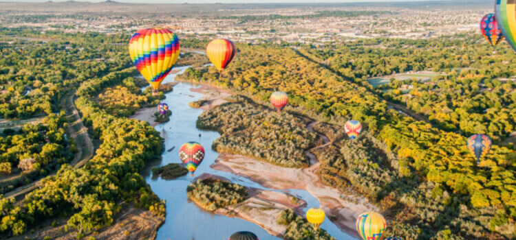 hot air balloons float above a river and forest with a new mexico city in the background