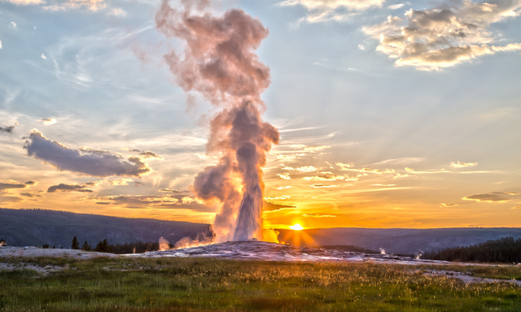 old faithful erupts with the sun setting behind the mountains