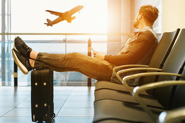 An airport passenger sits with their luggage and watches a plane take off outside a nearby window