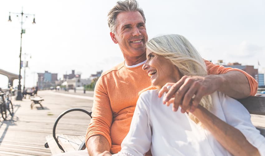 A mature couple smile and hold hands on a bench by the shire