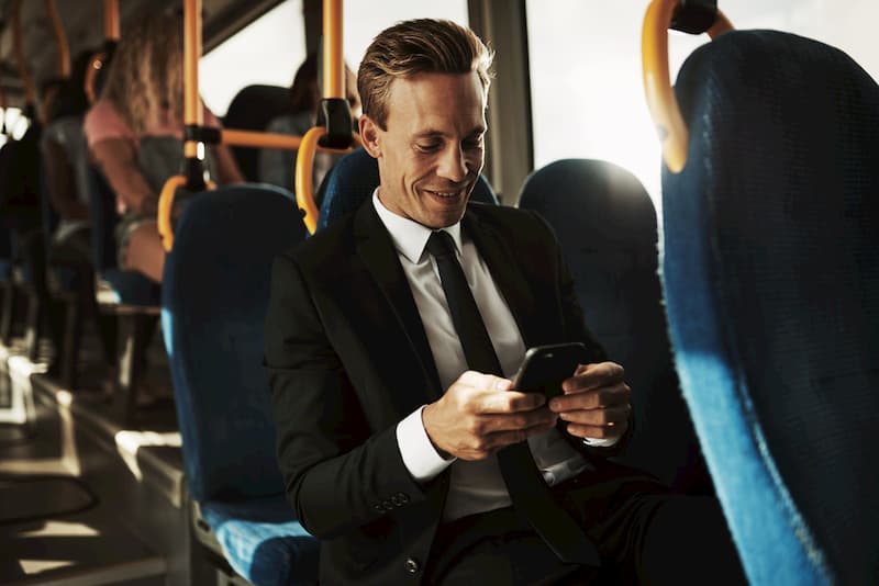 professional man in suit riding on charter bus looking at phone and smiling