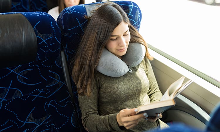 A woman with a neck pillow reads a book while riding on a charter bus
