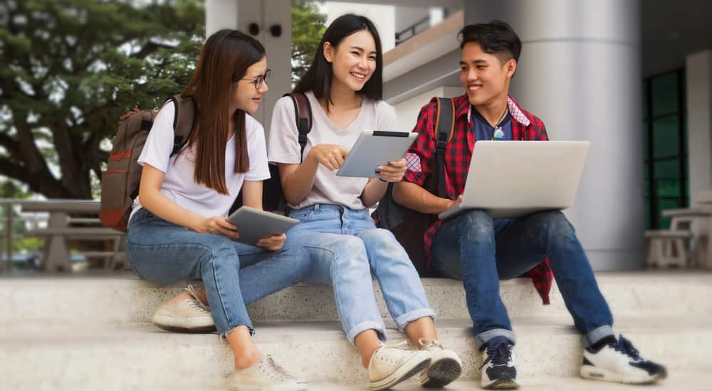 Three college students sit on stone steps of a building, smiling over their laptops