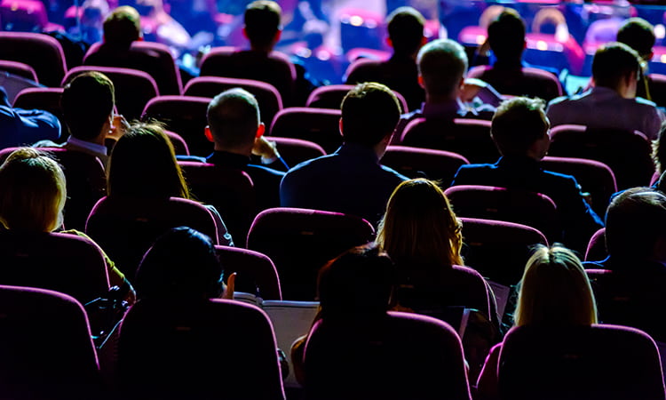 An audience sits in a dim room during a convention presentation