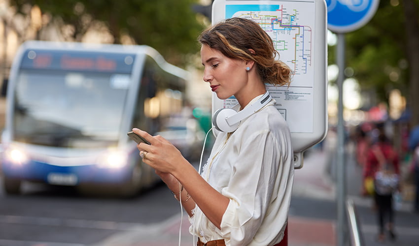 A college-aged student looks at her phone while waiting at a bus stop