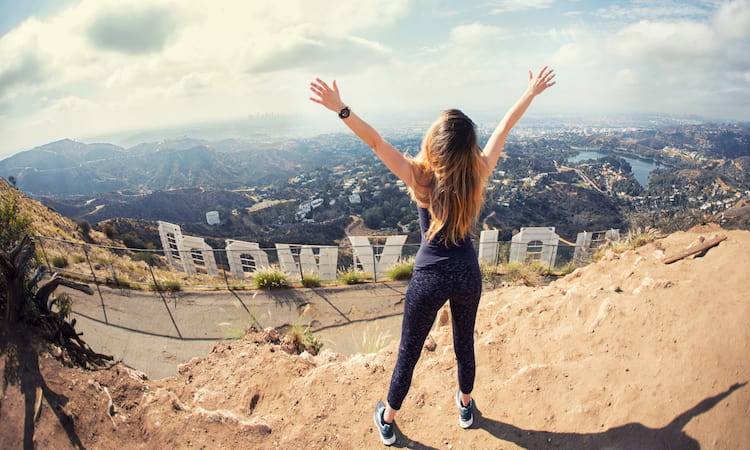 a woman hiker raises her arms to sky in triumph above the Hollywood sign