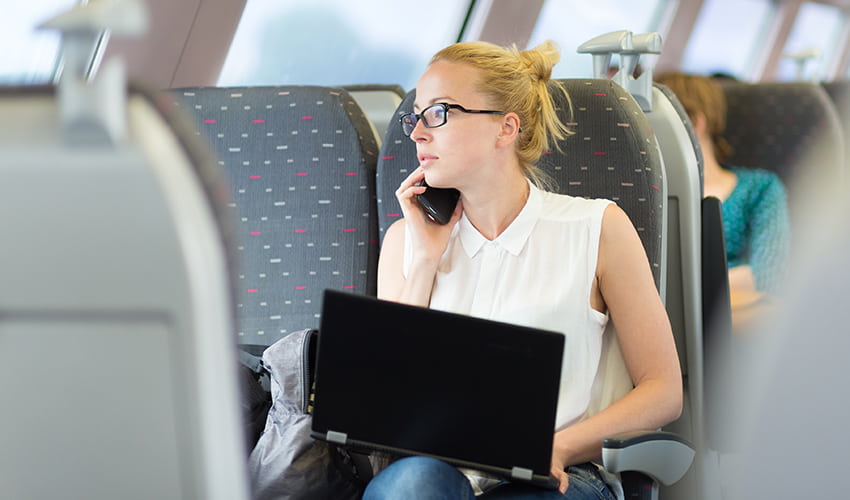 A woman on a commuter shuttle service types on a laptop while talking on a cell phone