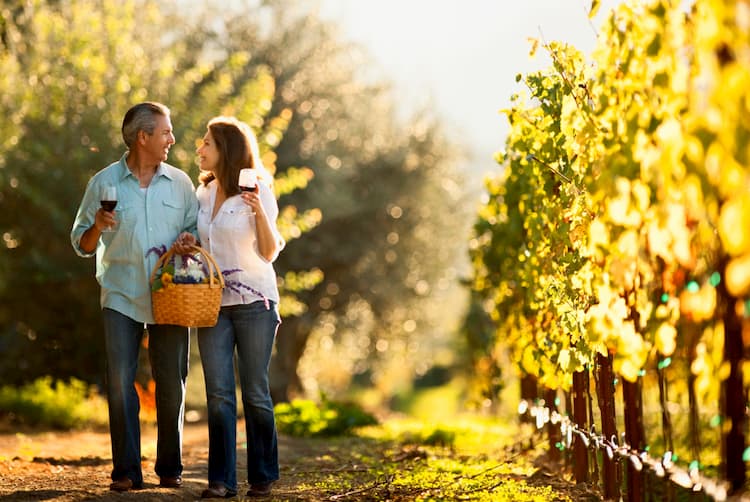 Couple walking in vineyard sipping glasses of wine.