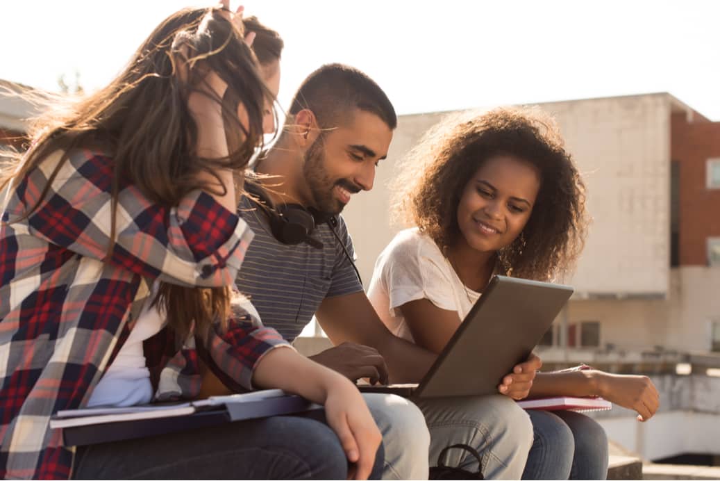 students look at a laptop and smile while outside on campus