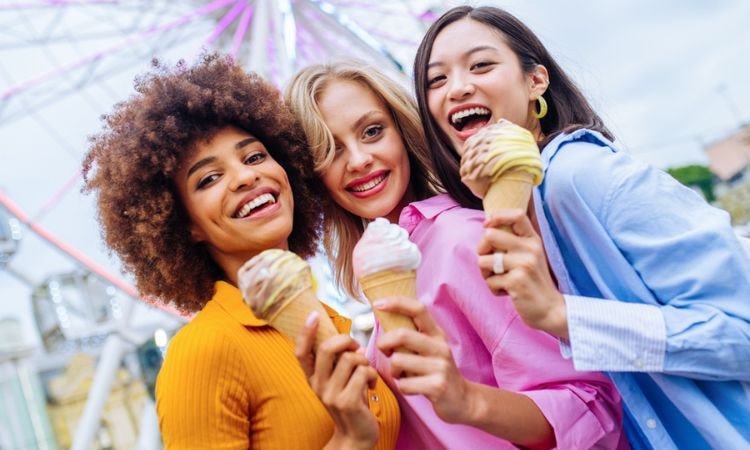 Three girls holding ice cream cones and smiling in front of a ferris wheel