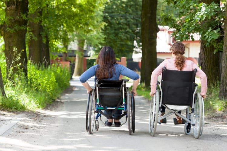 Two women in wheelchairs outdoors on a sidewalk in summer