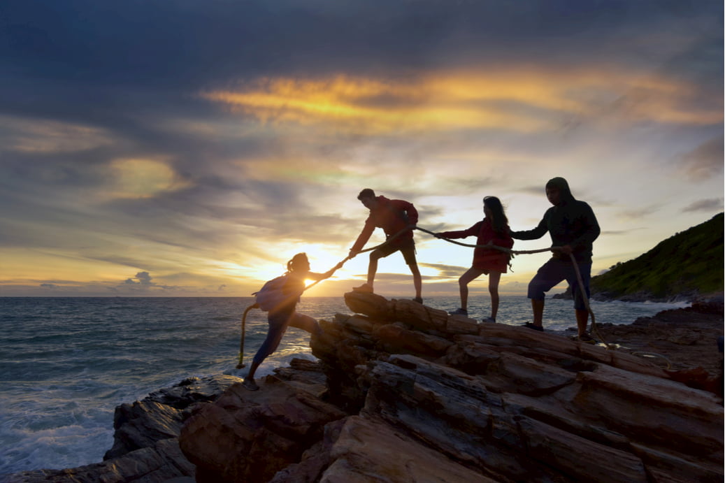 a group of friends help each other climb up the side of a rock, with a sunset in the background