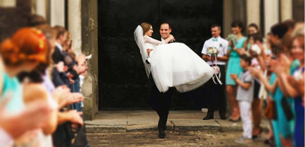 Bride being carried by groom with wedding guests clapping